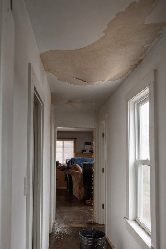 A water stain spreading across a hallway ceiling with a drip falling into a bucket